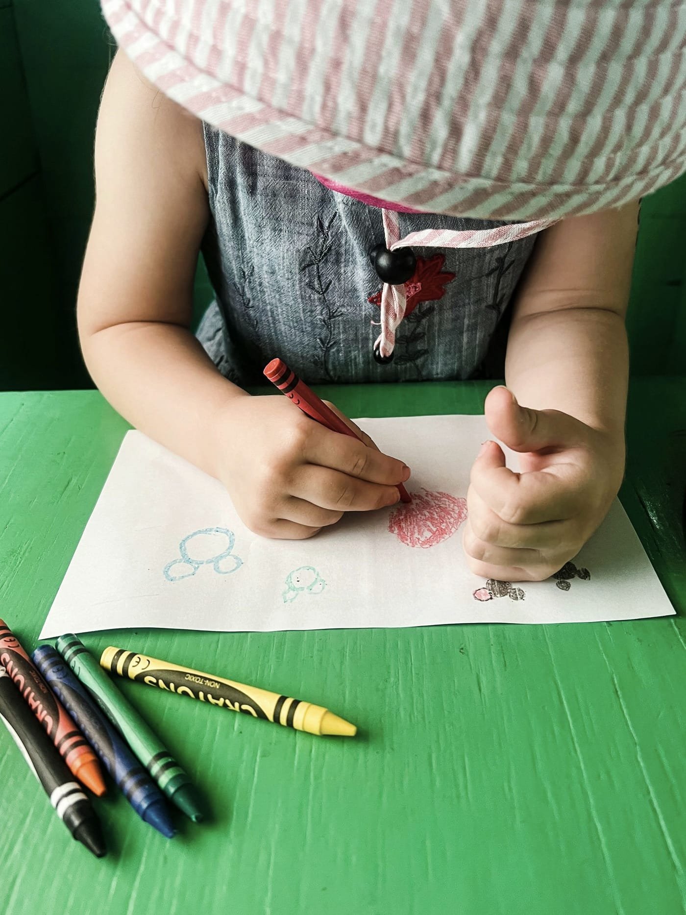 Mão de uma criança desenhando com giz de cera vermelho sobre uma folha de papel.