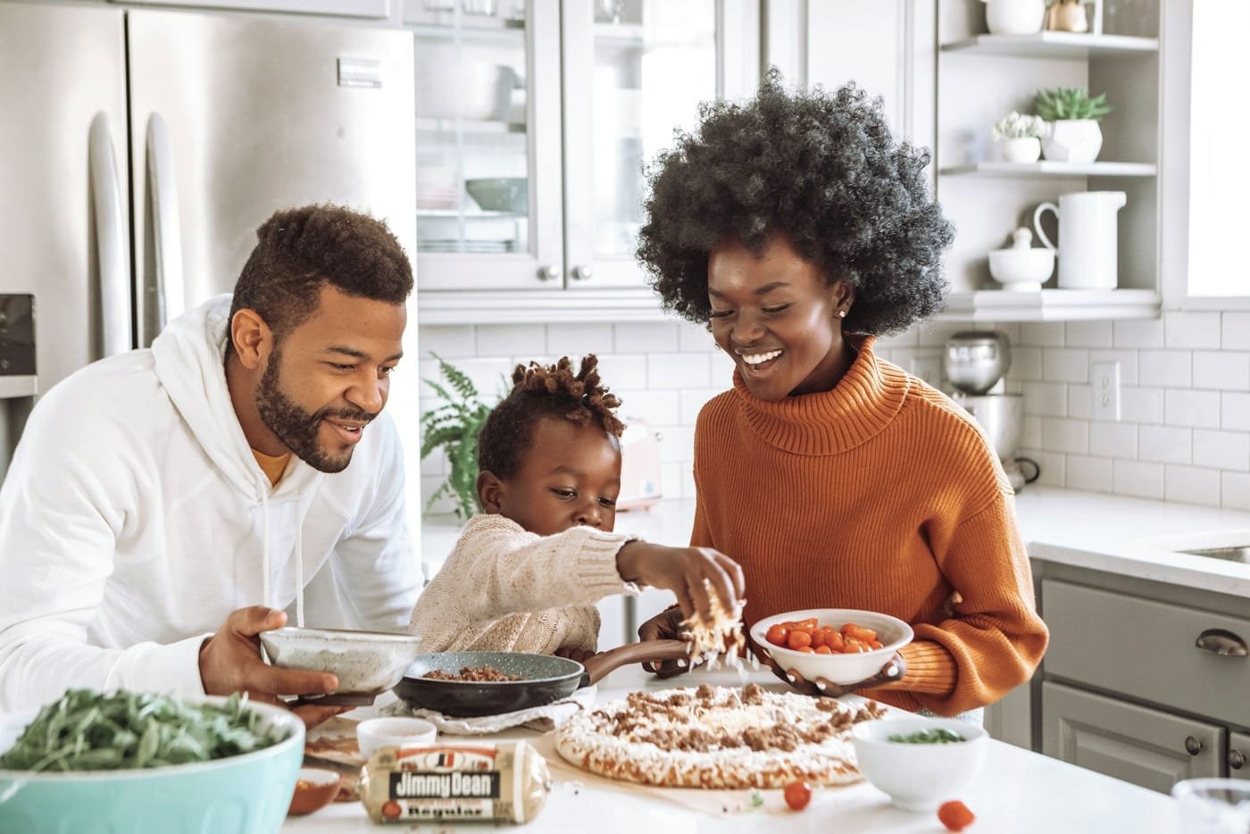 Pai, mãe e filho preparando uma pizza juntos na cozinha, sorrindo.